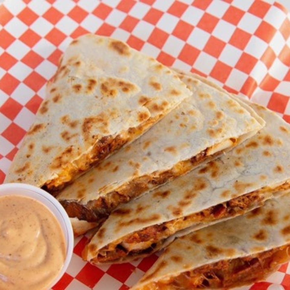Grilled quesadilla wedges filled with seasoned meat, served with creamy dipping sauce on red-and-white checkered paper.