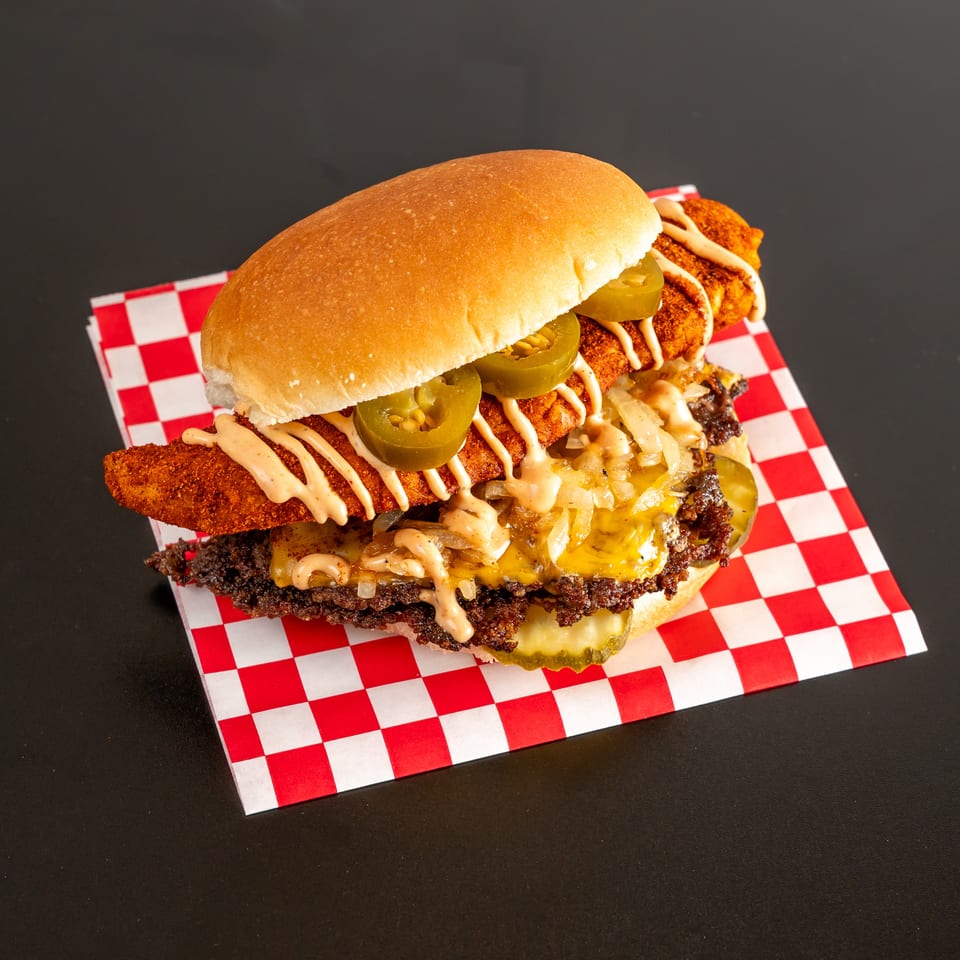 Loaded cheeseburger topped with a crispy fried fillet, jalapeños, grilled onions, creamy sauce, and pickles on checkered paper.