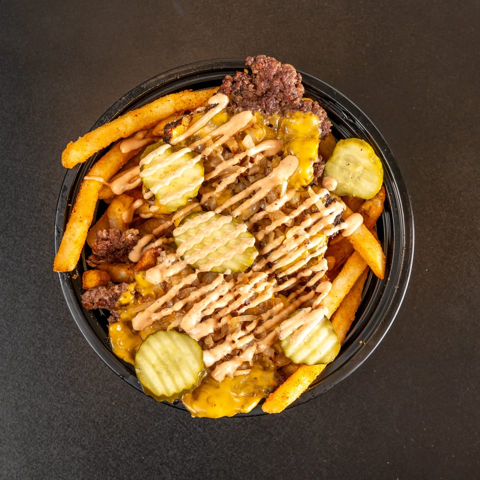 Loaded fries topped with cheeseburger pieces, melted cheddar, pickles, grilled onions, and drizzled sauce in a black bowl.