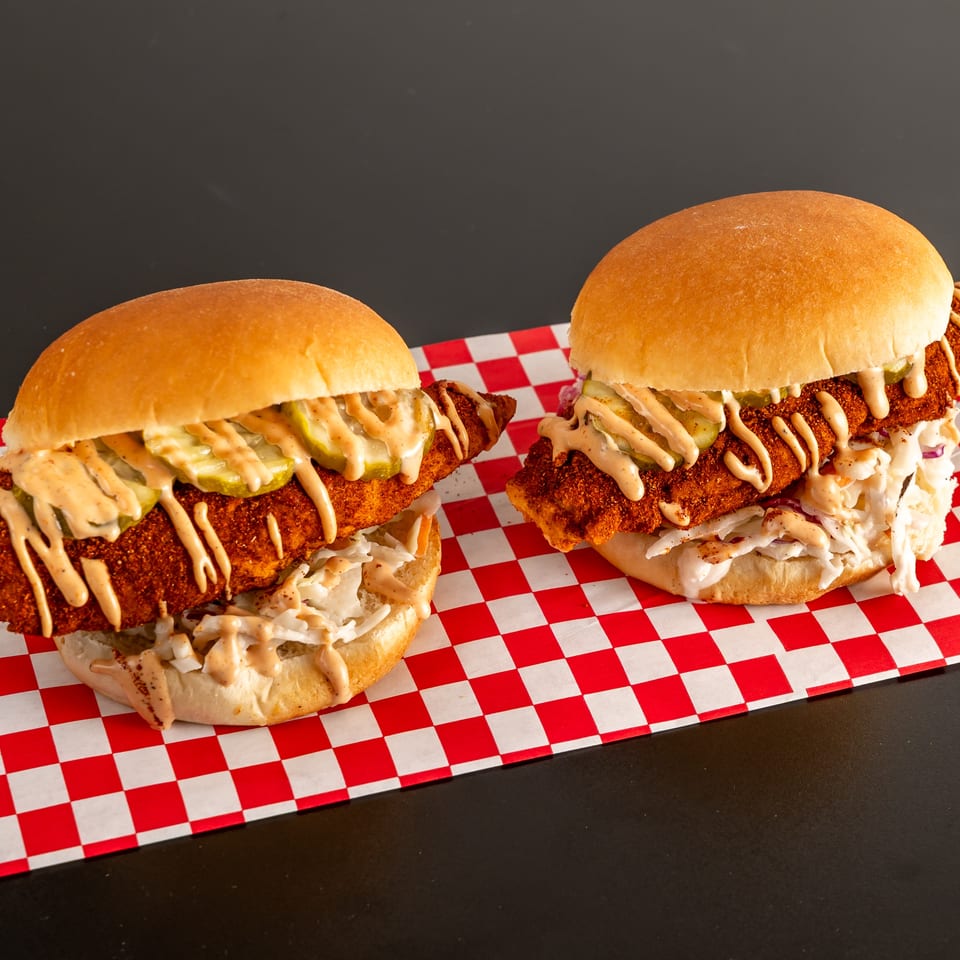 Two crispy fried chicken sandwiches with pickles, creamy slaw, and drizzled sauce on red-and-white checkered paper.