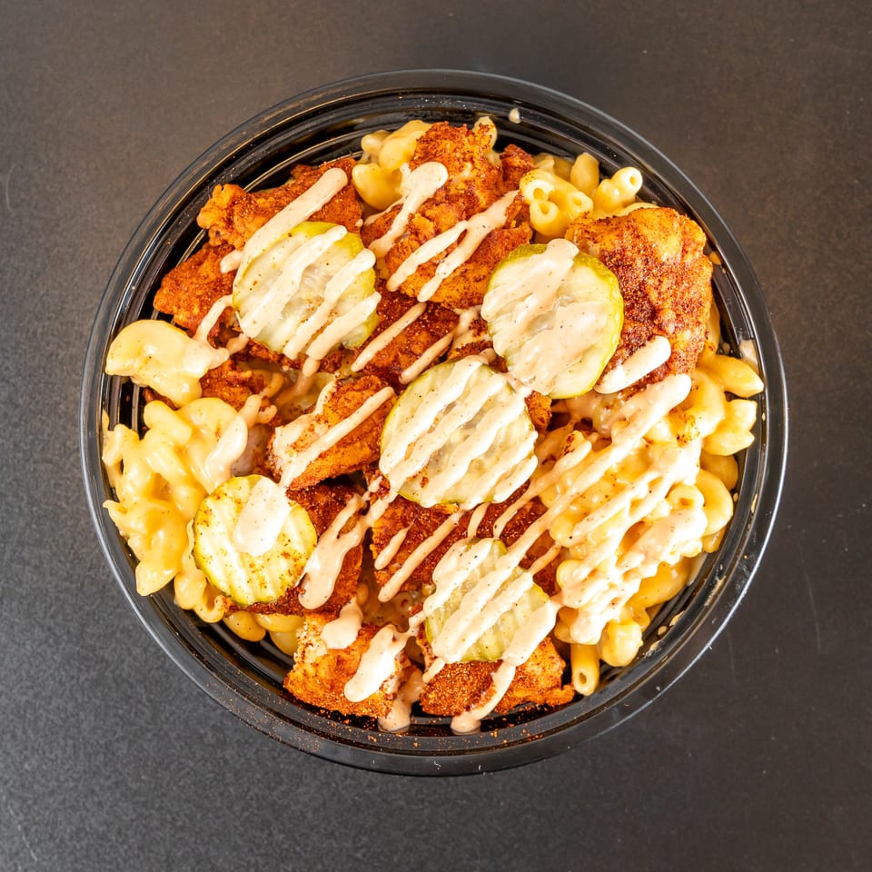 Bowl of seasoned fried chicken over creamy macaroni and cheese, topped with pickles and drizzled sauce.