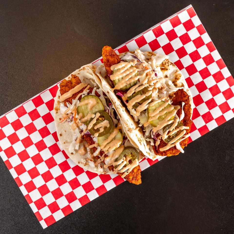 Three crispy fried chicken tacos topped with coleslaw, pickles, and creamy sauce on red-and-white checkered paper.