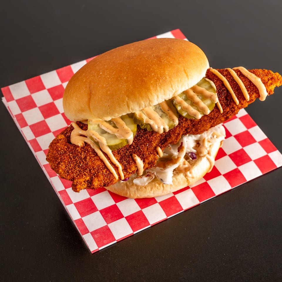 Crispy breaded chicken sandwich with pickles, creamy sauce, and coleslaw on red-and-white checkered paper.