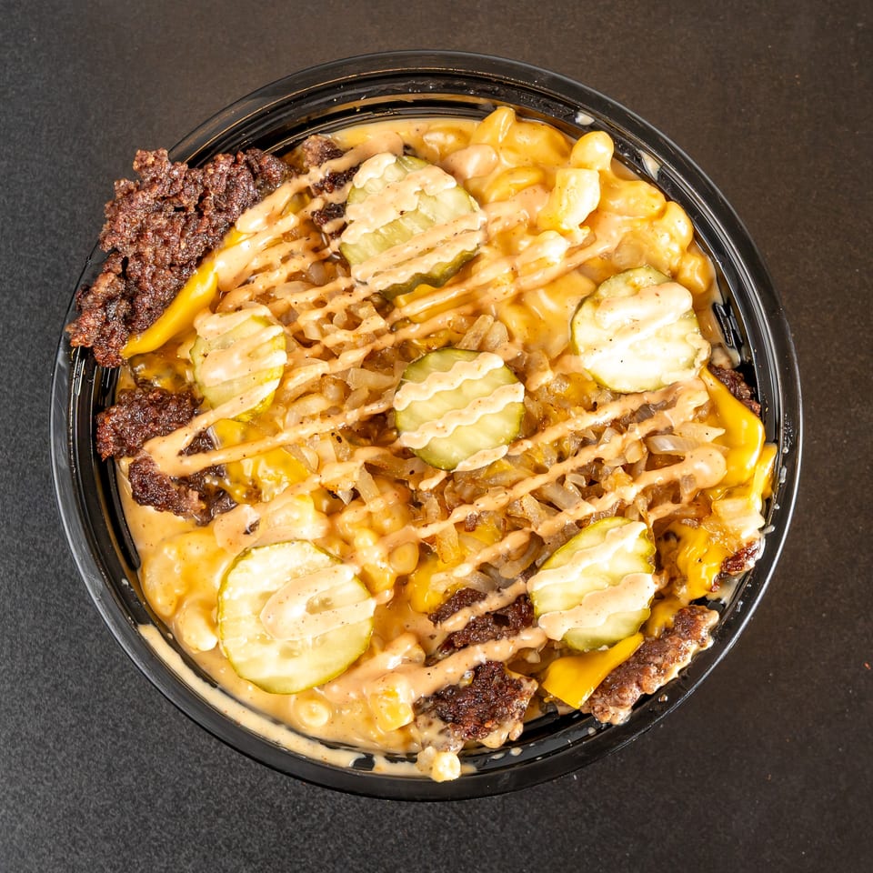 Loaded bowl of cheeseburger macaroni topped with grilled beef, pickles, onions, melted cheese, and drizzled sauce.