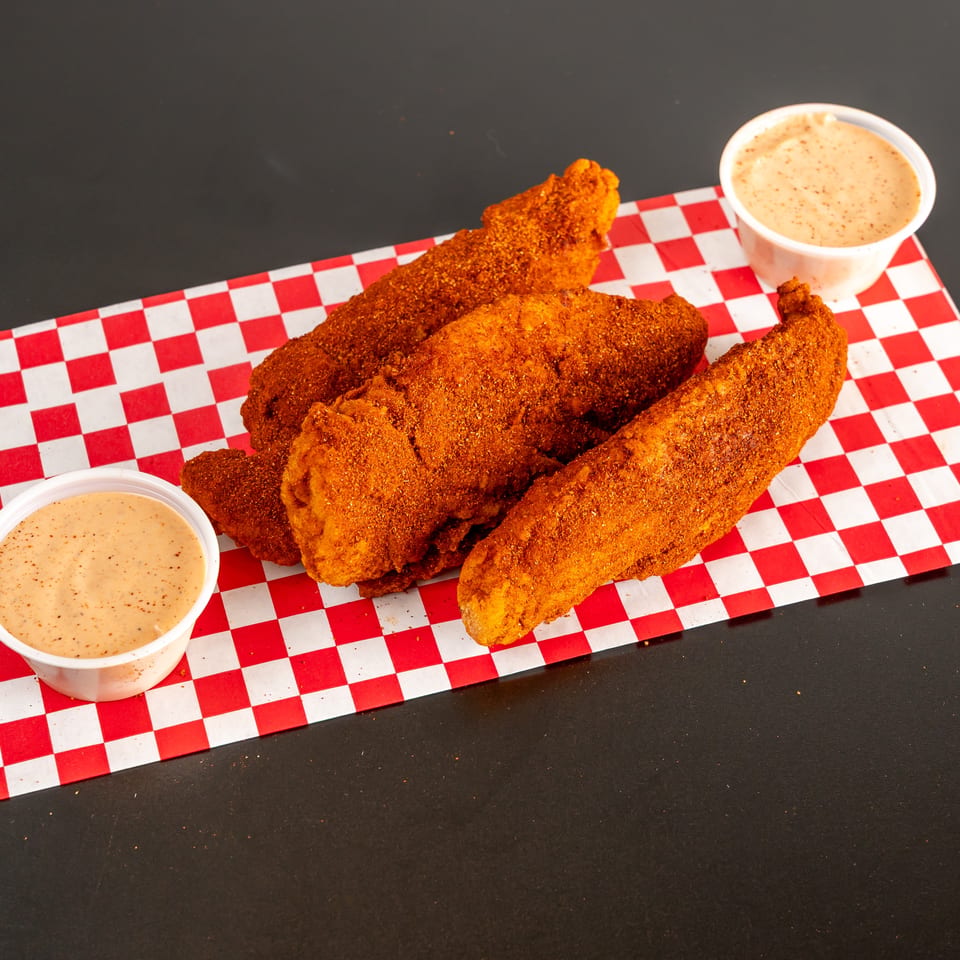 Crispy seasoned fried chicken tenders arranged on red-and-white checkered paper with two cups of creamy dipping sauce.
